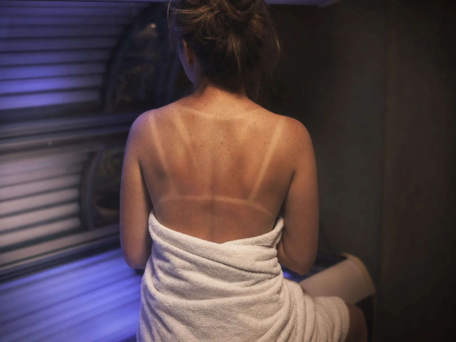 A woman in her thirties sitting on the edge of an open tanning bed, seen from behind, with strap tan lines across her shoulders