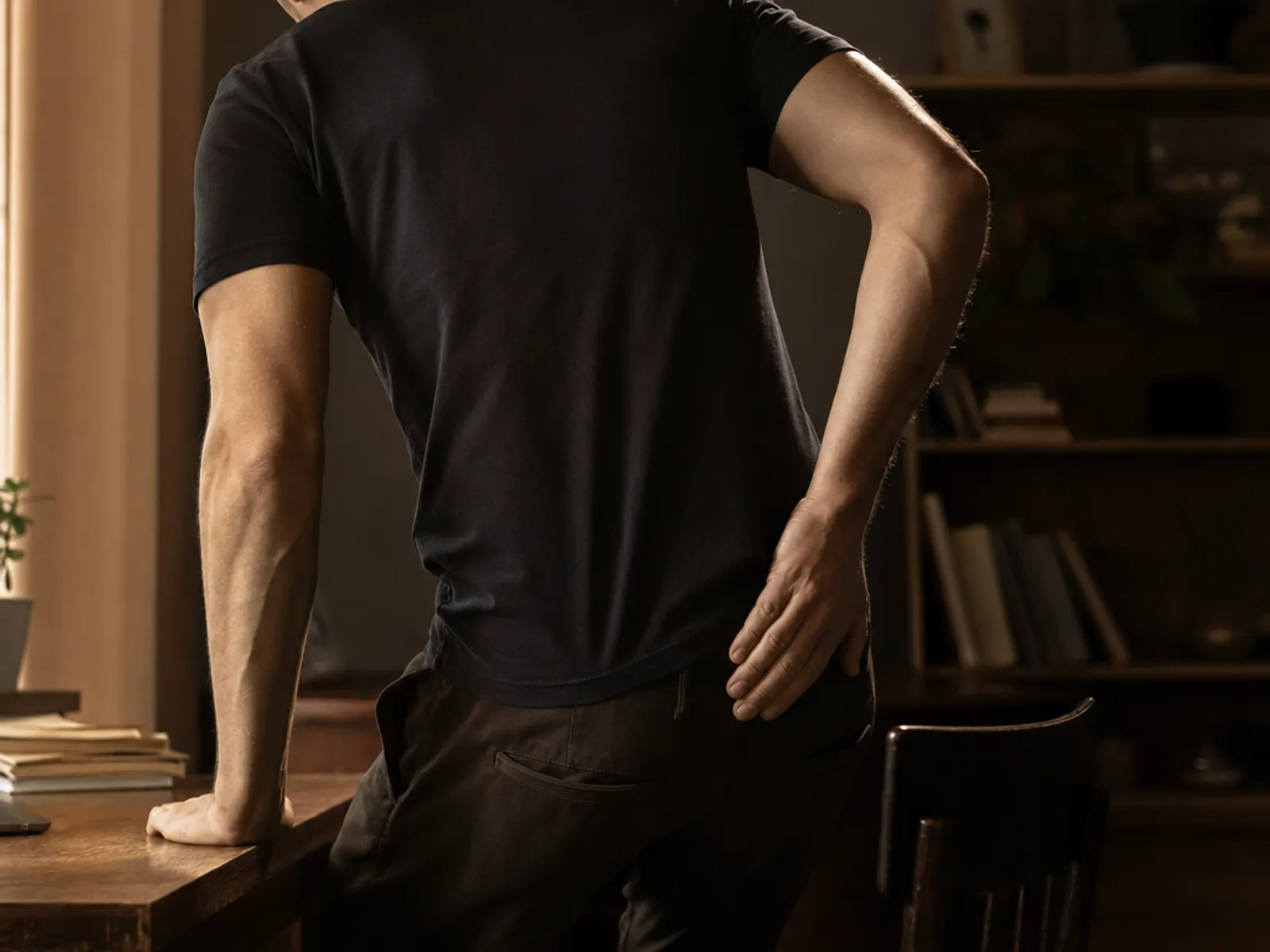 A young man seen from behind, pausing as he sits down at a desk with one hand pressed to his lower back
