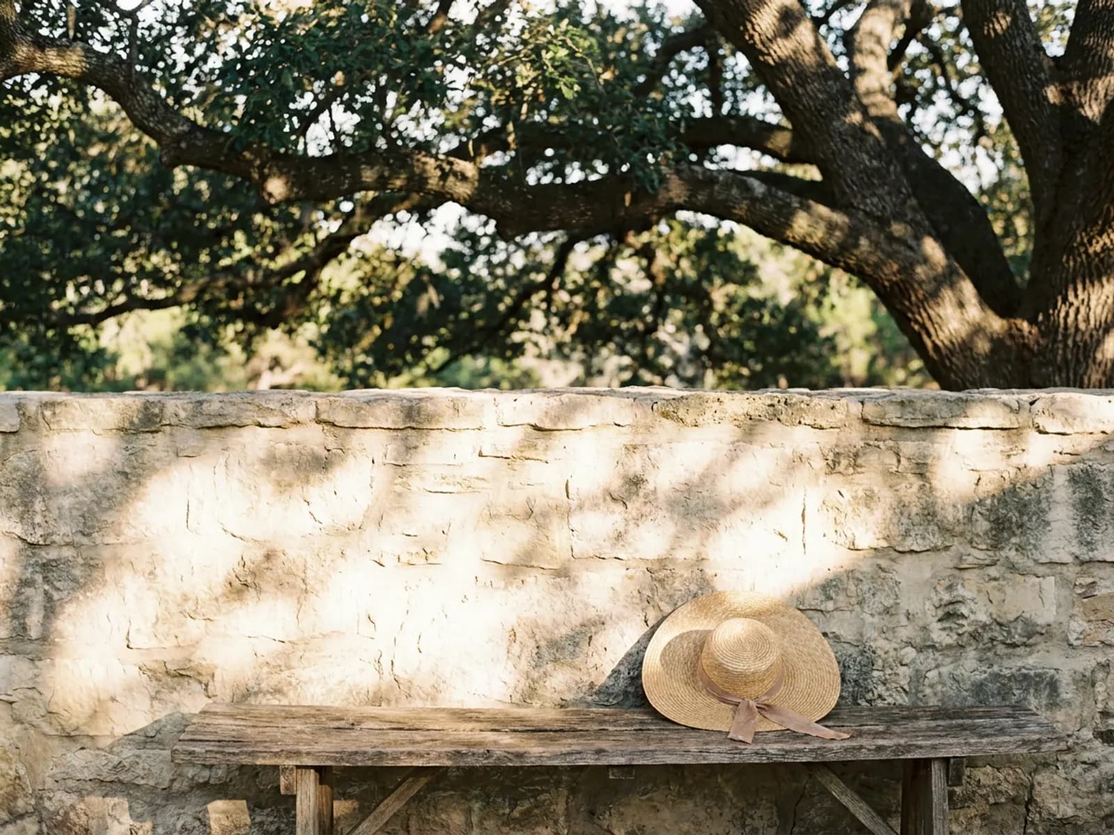 Dappled afternoon sunlight through a live oak falling on a limestone wall and a straw hat on a wooden bench
