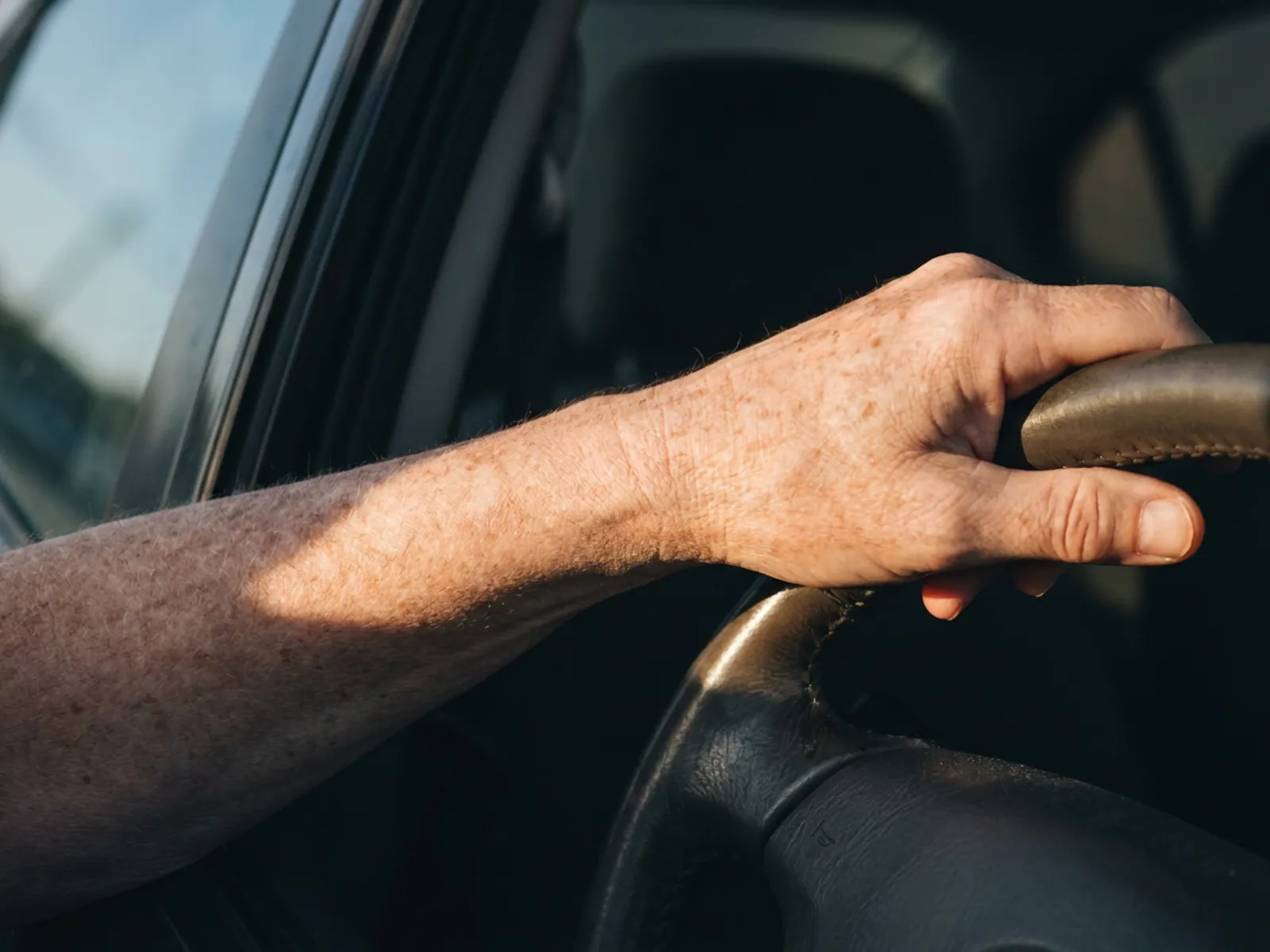 A driver's left forearm on the steering wheel with a hard band of sunlight falling across it through the closed side window